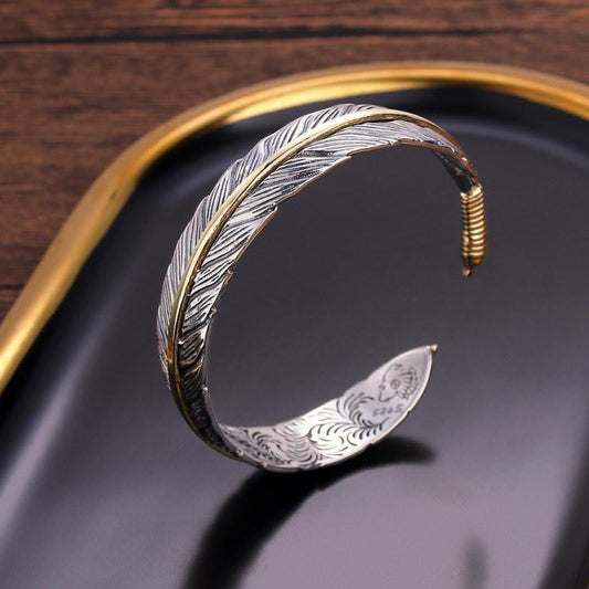 Two-tone silver and gold feather cuff bracelet with etched barb details, displayed on black tray with gold rim against wood background