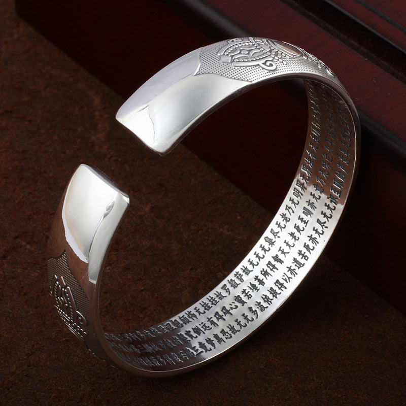 Tibetan silver prayer bangle with circular medallion motifs, lotus and dharma wheel patterns, Heart Sutra Chinese text interior on dark surface.