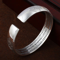 Tibetan silver prayer bangle with circular medallion motifs, lotus and dharma wheel patterns, Heart Sutra Chinese text interior on dark surface.