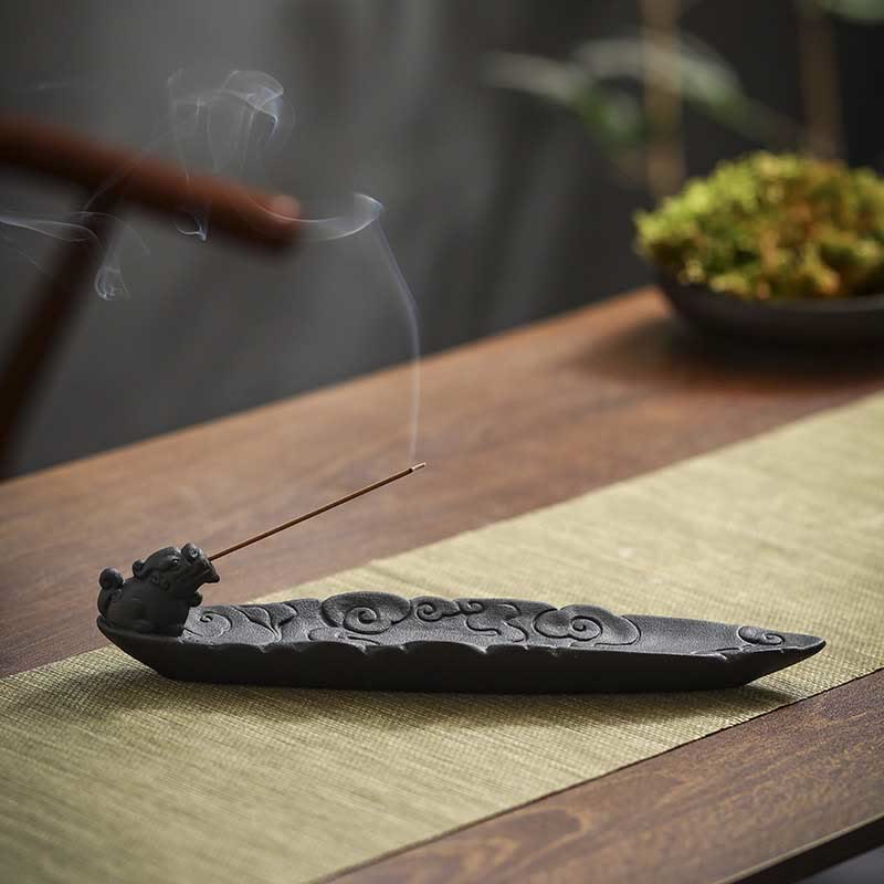 Low-angle Pixiu incense holder with wispy smoke, woven mat, bamboo bokeh, green plant accents – dark ceramic incense burner