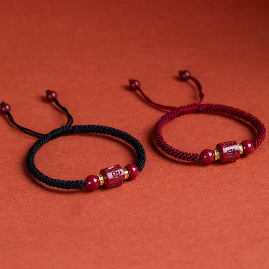 Pair of adjustable braided cord bracelets in burgundy red and black with carved cinnabar Tibetan mantra cylinder beads, gold spacer rings and polished red accent beads on rust background