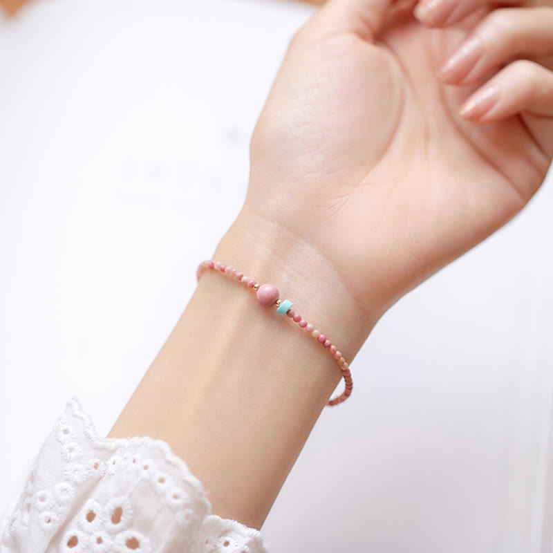 Rhodonite Loving Energy Bracelet – Pink beads with rose focal and turquoise accent worn on wrist, soft white bokeh background with faint pink shapes