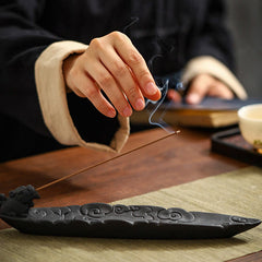 Hand reaching toward smoke from Pixiu incense holder, tea cup nearby, woven olive mat, dark ceramic – auspicious cloud incense burner