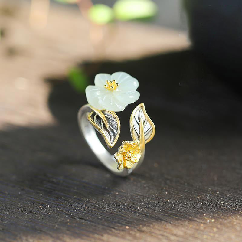 White Jade Plum Blossom Silver Ring - Floral ring against green foliage with carved jade flower, gold and silver two-tone metalwork, and oxidized leaf details