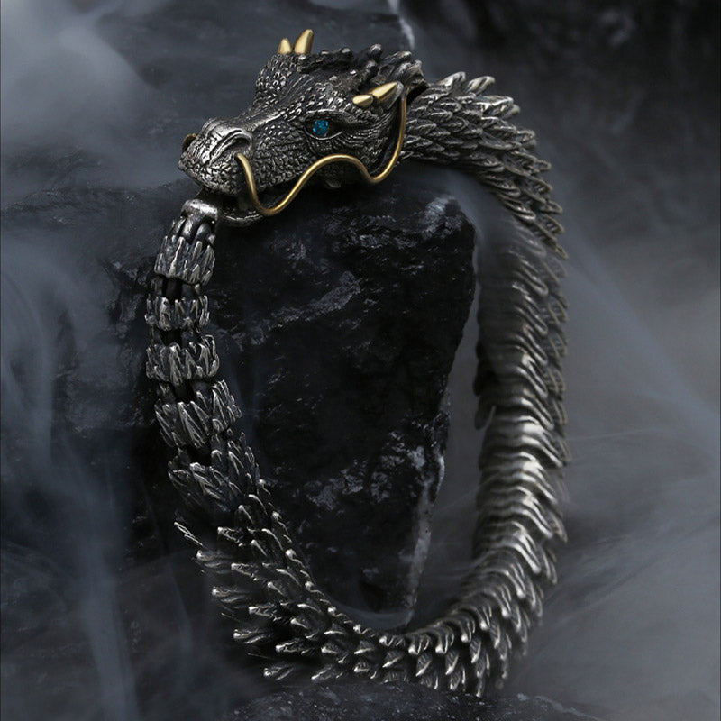 Dragon-themed articulated cuff bracelet with oxidized silver body gold whiskers and horns blue gemstone eye displayed on volcanic rock with dramatic smoke effects