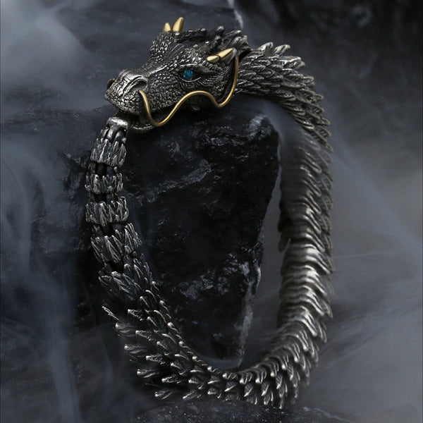 Dragon-themed articulated cuff bracelet with oxidized silver body gold whiskers and horns blue gemstone eye displayed on volcanic rock with dramatic smoke effects