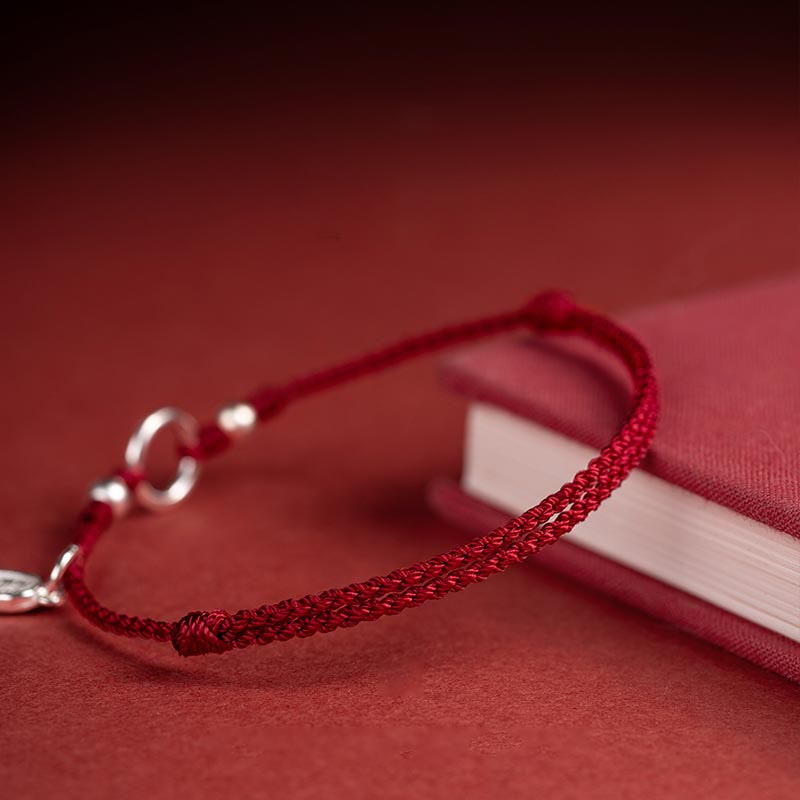 Sterling Silver Lotus Peace Buckle Red String Bracelet – Angled close-up of red braided cord with silver ring clasp and cylindrical beads alongside a red book prop