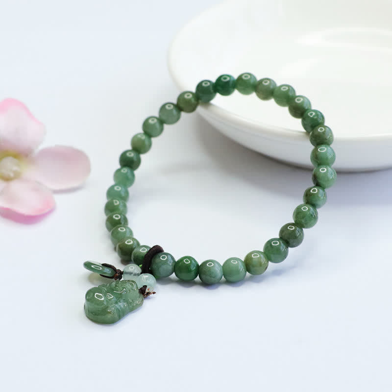 Natural green jade stretch bracelet with carved jade Pixiu mythical creature charm and translucent accent beads on white background with pink flower decoration