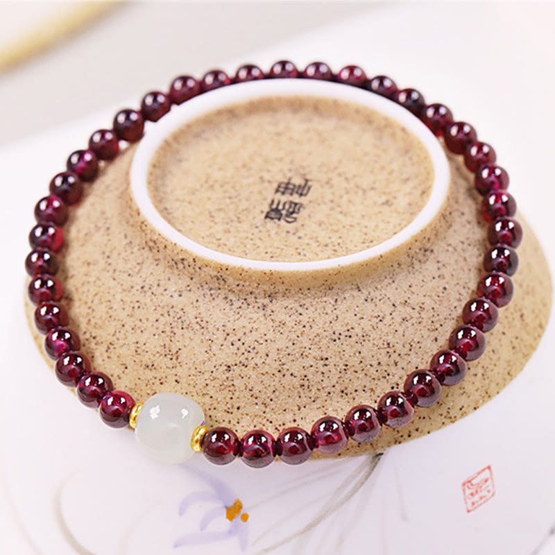Garnet and white jade bracelet displayed on antique ceramic teacup with calligraphy, showing burgundy beads with jade center bead and gold accents