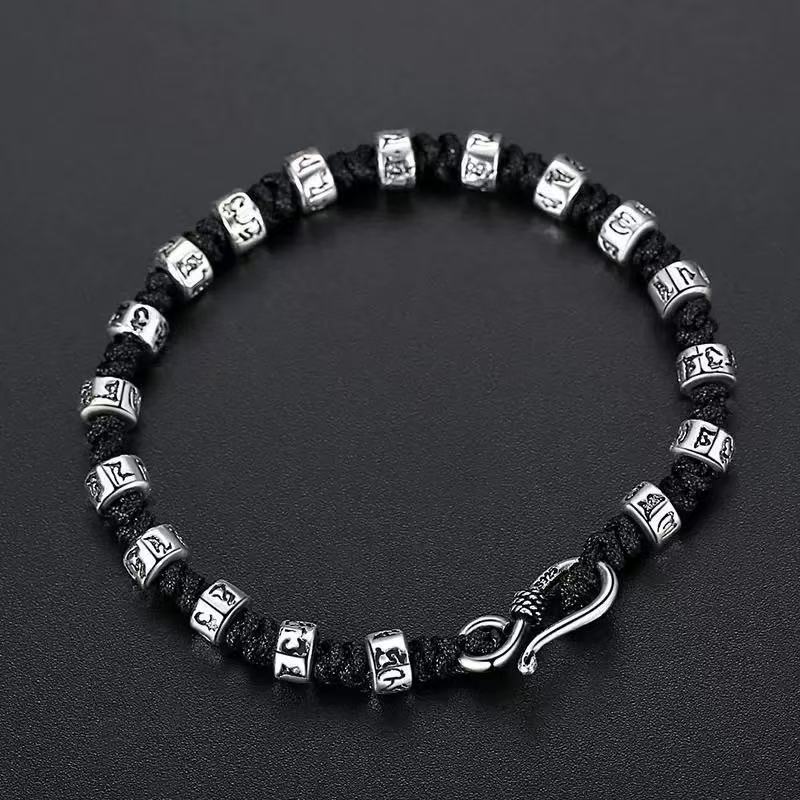 Zen Mantra Cord Bracelet – overhead view of silver Tibetan mantra cylinder beads and curved S-clasp on black cord, dark background