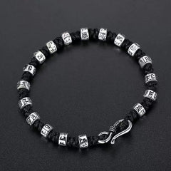 Zen Mantra Cord Bracelet – overhead view of silver Tibetan mantra cylinder beads and curved S-clasp on black cord, dark background