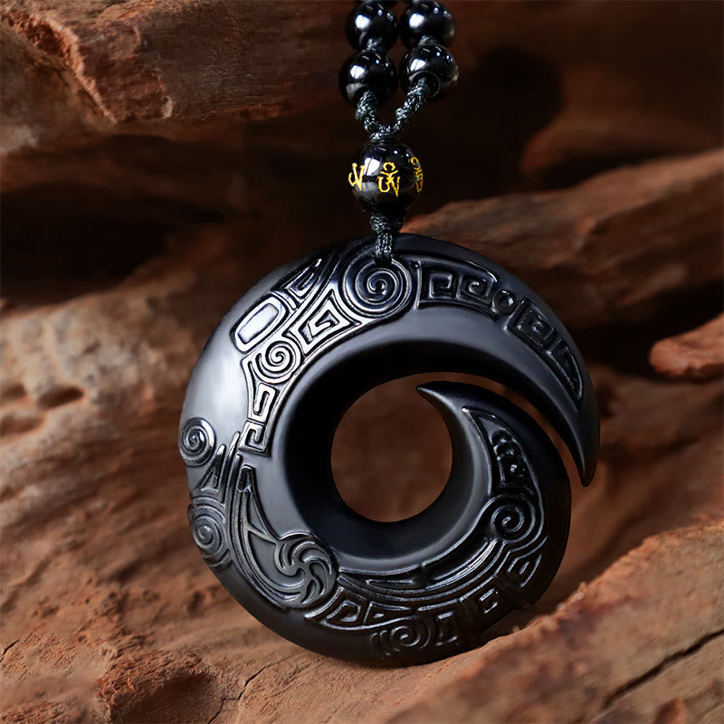Black Obsidian Ice Obsidian Pendant Necklace – Black obsidian spiral vortex pendant with detailed relief carving on natural wood and stone background, gold Sanskrit character accent bead, earthy artisanal product photography, grounding energy crystal necklace