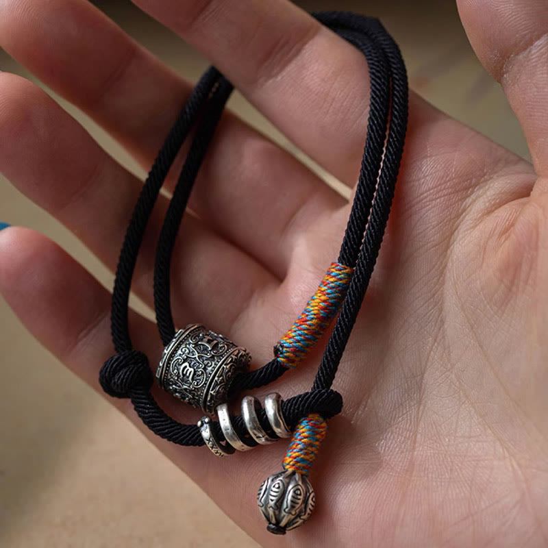 Tibetan Om Mani Padme Hum Wrap Bracelet – Silver mantra bead, multiple rings and fish-tail charm on black cord, beige fabric angled view