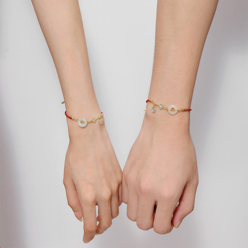Pair of matching red cord bracelets with pale green jade peace buckle discs and gold-toned charms worn on two wrists against white background