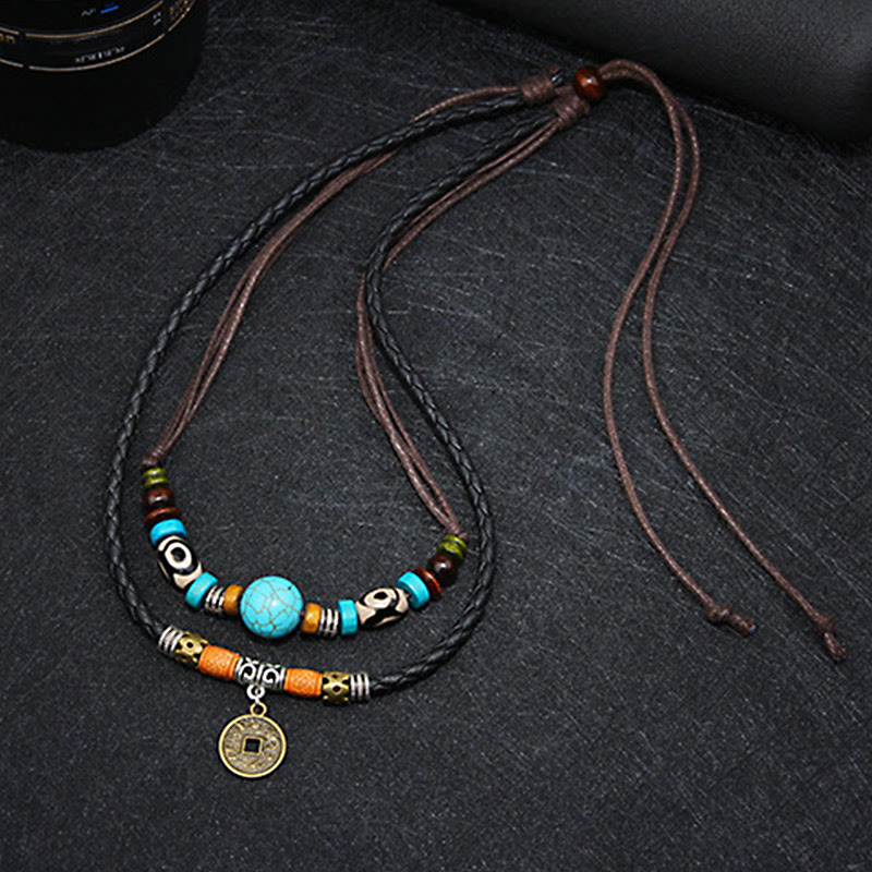 Turquoise Dzi Bead Protection Necklace – top-down flat-lay on dark marbled surface showing complete double-strand design with turquoise bead, Dzi eye beads, orange amber accents, and bronze coin charm, brown braided leather cord with adjustable sliding knot