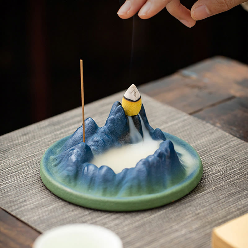 Ceramic Backflow Incense Burner – Hand placing cone on blue mountain, smoke cascading on gray mat, meditation