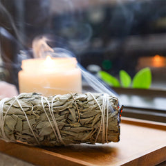 Burning white sage smudge stick with smoke on wooden tray, lit candle nearby