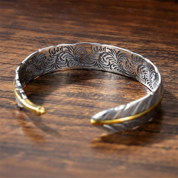 Sterling silver feather motif cuff bracelet with gold accents, detailed barb texture and ornate interior floral engraving on wood surface
