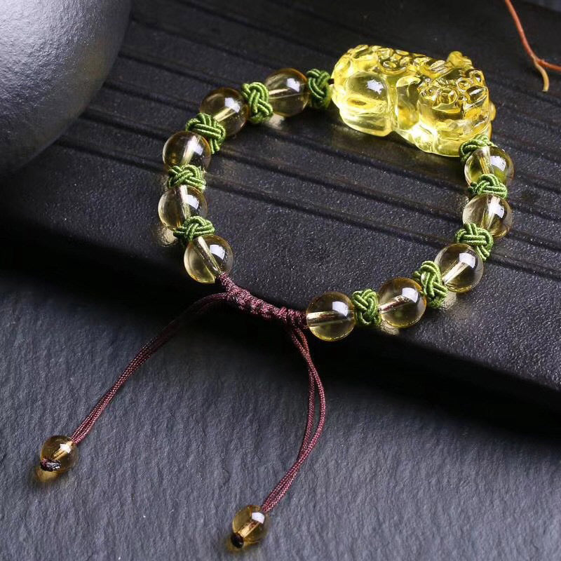 Natural Citrine PiXiu Wealth Bracelet – overhead three-quarter view showing golden amber citrine beads, detailed PiXiu carving, green knotwork between beads, and burgundy sliding knot closure, on dark textured stone surface with ceramic prop