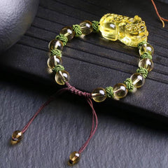 Natural Citrine PiXiu Wealth Bracelet – overhead three-quarter view showing golden amber citrine beads, detailed PiXiu carving, green knotwork between beads, and burgundy sliding knot closure, on dark textured stone surface with ceramic prop