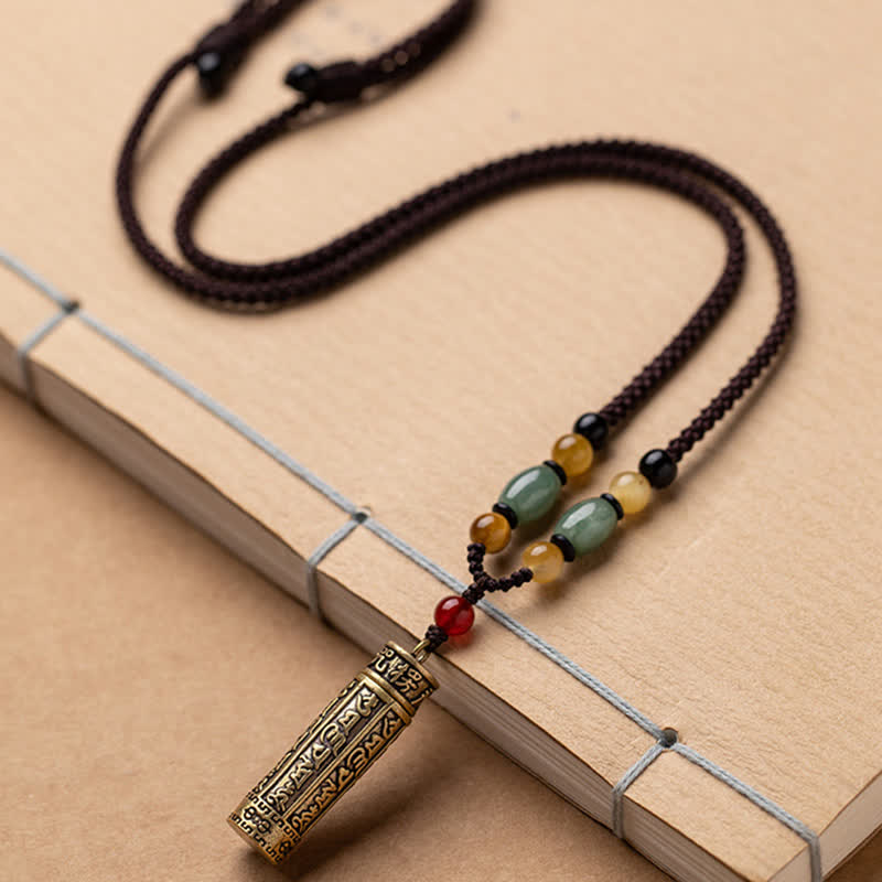 Vintage brass cylinder pendant necklace with engraved symbols and green jade barrel beads, amber accents, adjustable brown cord on Chinese book