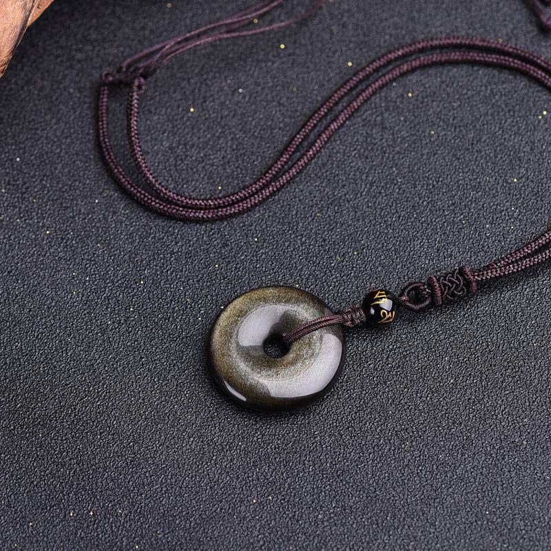 Golden bronze sheen obsidian peace buckle pendant necklace with gold Tibetan script bead on burgundy braided cord on dark textured fabric with subtle gold glitter accents