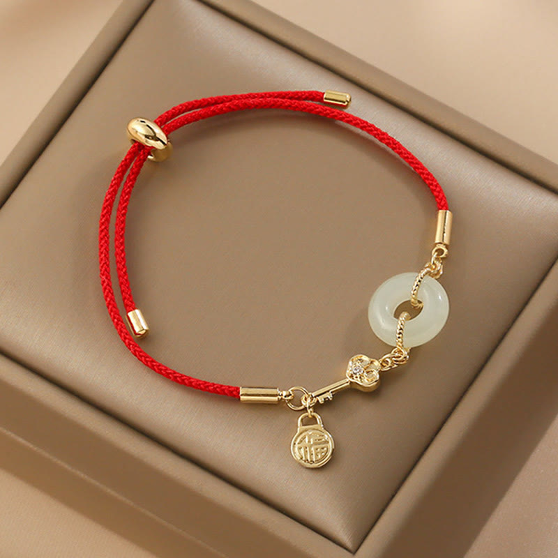 Chinese red cord bracelet with pale green jade peace buckle, gold key charm with crystal accent and fortune lock pendant in beige velvet jewelry box