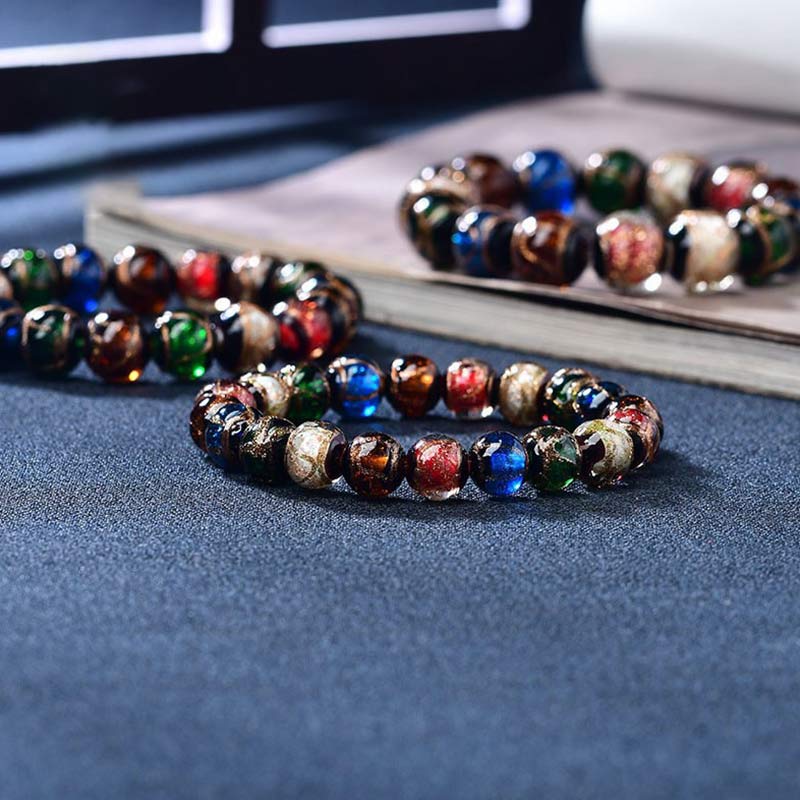 Colorful Liuli Glass Bead Luck Bracelet – Multicolored iridescent glass beads with blue green amber hues on cream fabric display surface with gold stand, low side
