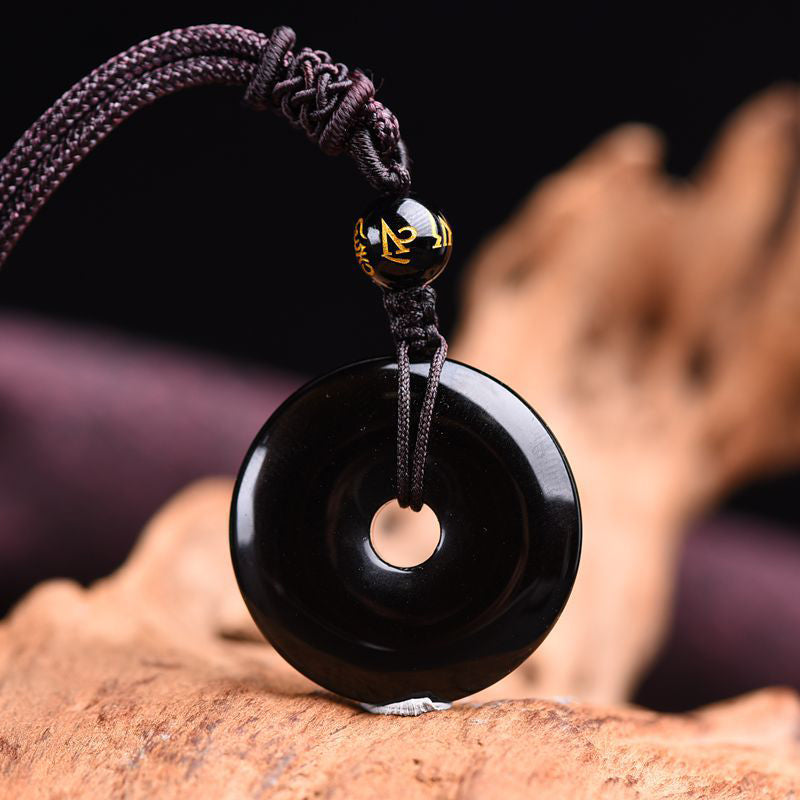 Black obsidian ping'an kou peace buckle pendant necklace with gold-inscribed Tibetan mantra bead on dark braided cord displayed on natural weathered driftwood