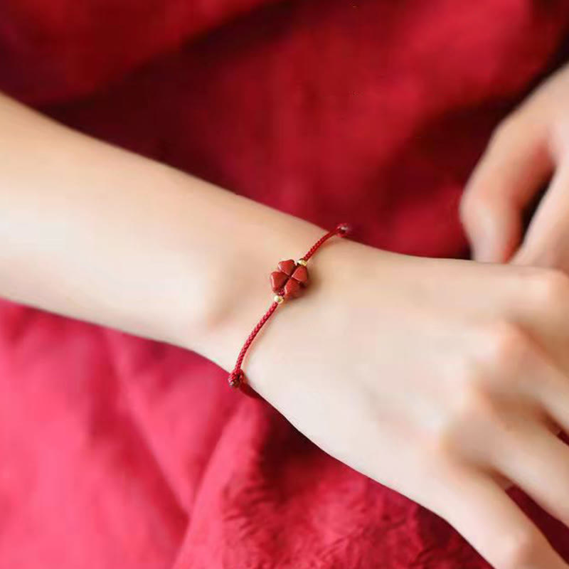 Cinnabar four leaf clover red cord bracelet worn on wrist showing carved vermilion charm gold accent beads against draped red silk fabric lifestyle model shot