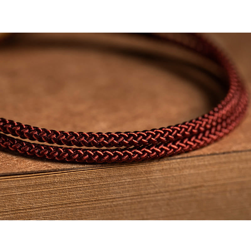 925 Silver Zodiac Natal Buddha Red String Bracelet detail shot on book pages, burgundy braided cord with natural drape, minimalist artisanal styling, warm brown tones
