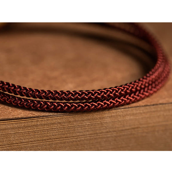 925 Silver Zodiac Natal Buddha Red String Bracelet detail shot on book pages, burgundy braided cord with natural drape, minimalist artisanal styling, warm brown tones