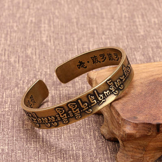 Antique brass Tibetan mantra cuff bracelet with embossed Sanskrit script exterior and Chinese characters inside, displayed on wooden stand against woven fabric background