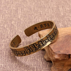 Antique brass Tibetan mantra cuff bracelet with embossed Sanskrit script exterior and Chinese characters inside, displayed on wooden stand against woven fabric background