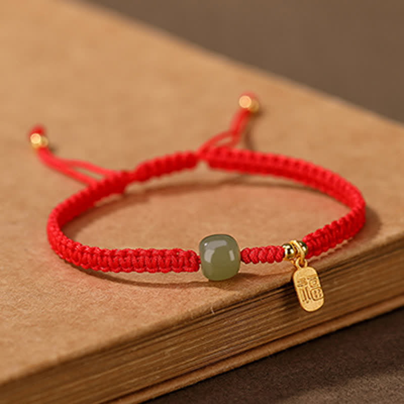 Red cord bracelet with Hetian jade bead gold spacer beads fu fortune charm and gold cord end caps on stacked vintage books display with artisanal lifestyle aesthetic