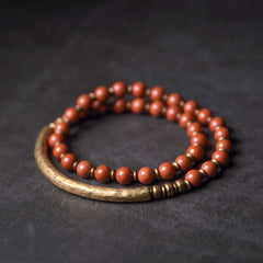 Red jasper wrap bracelet with antique brass curved bar and hammered metal spacer beads on dark slate background, bohemian artisan double-coil design