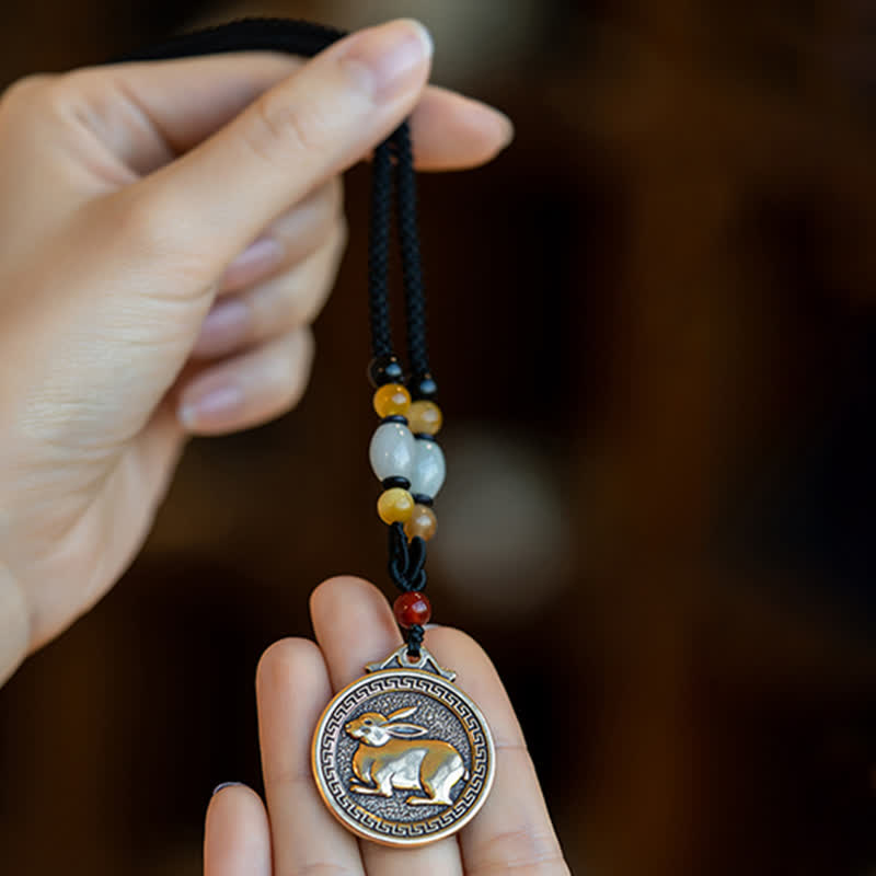 12 Chinese Zodiac Fortune Necklace Pendant – Rabbit zodiac pendant necklace held in hand, two-tone silver and rose gold medallion with rabbit relief and meander border, multi-color jade amber beads on black cord, warm bokeh background, zodiac birth year jewelry