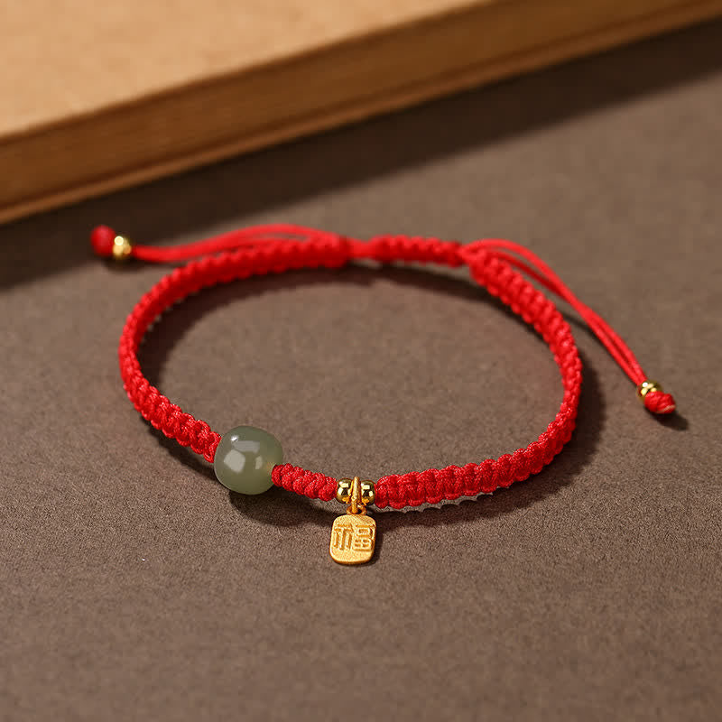 Adjustable red cord bracelet with Hetian jade green bead gold fu blessing fortune charm and sliding knot closure on neutral brown textured background with wooden prop accent