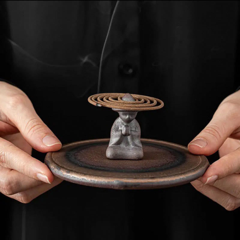 Hands holding gray monk figure with coil incense on brown base, healing