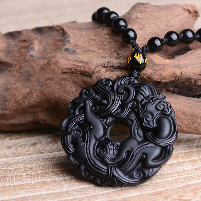 Black Obsidian Pixiu Pendant – Carved obsidian peace buckle resting on driftwood, beaded cord