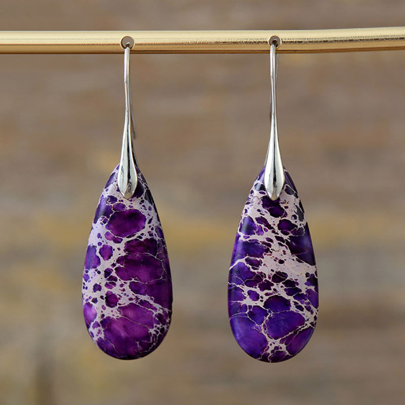Emperor Stone Healing Crystal Earrings – Purple teardrop charoite stone earrings with white swirling veins and silver ear hooks on brass display rod