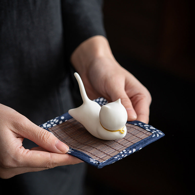 Hands holding white ceramic cat figurine with gold collar on bamboo floral mat