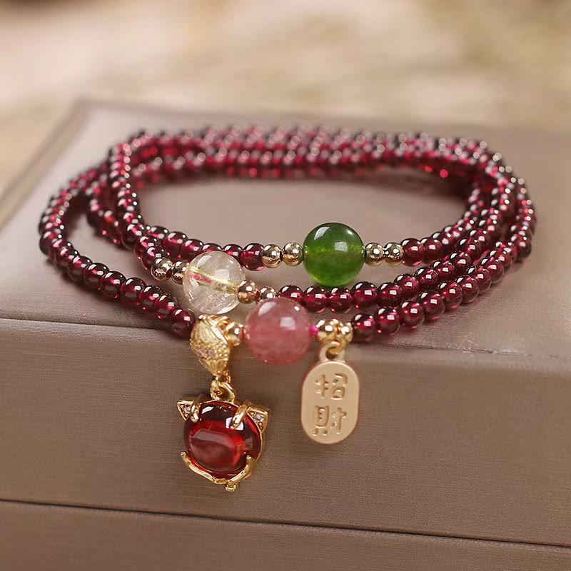 Triple-strand garnet bracelet with fox face charm red cabochon gemstone zhao cai wealth invitation gold tag and mixed semi-precious accent beads on taupe display surface