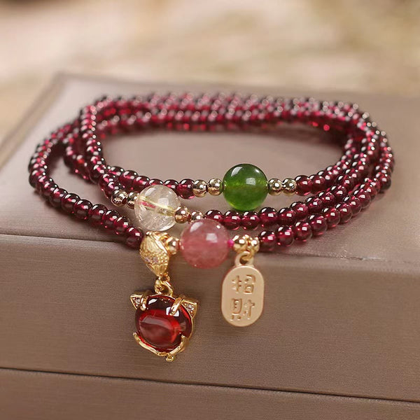 Triple-strand garnet bracelet with fox face charm red cabochon gemstone zhao cai wealth invitation gold tag and mixed semi-precious accent beads on taupe display surface