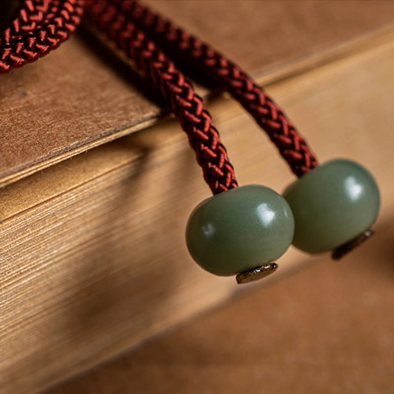925 Silver Zodiac Natal Buddha Red String Bracelet close-up on aged paper, two celadon jade barrel beads with cord texture detail, burgundy braided cord macro photography showing craftsmanship