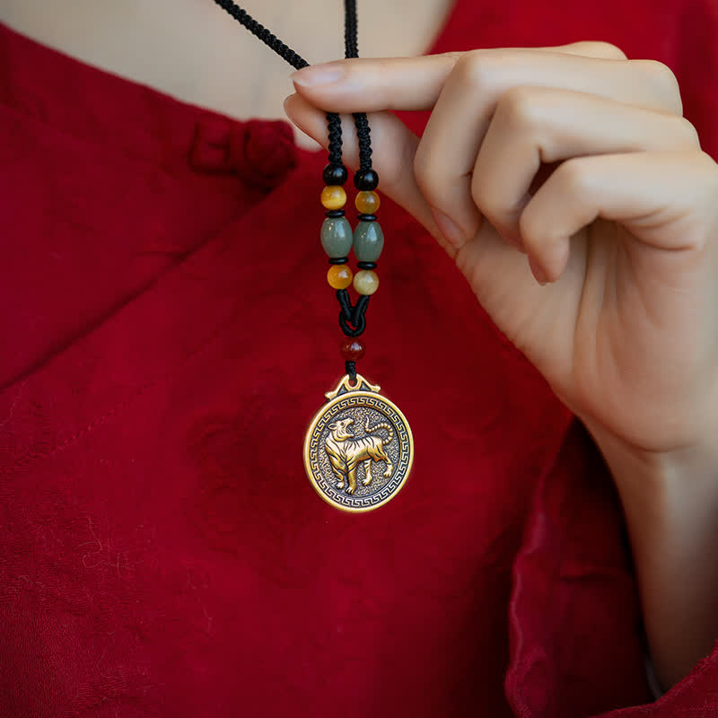 12 Chinese Zodiac Fortune Necklace Pendant – Close-up of tiger zodiac pendant on red fabric, gold tone medallion with walking tiger relief and Greek key pattern border, hand grasping beaded cord, studio lighting, Chinese zodiac protective talisman