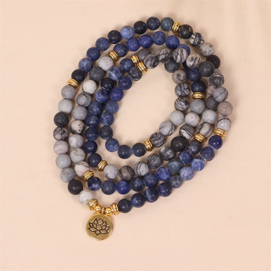 Triple-strand sodalite and zebra jasper mala wrap bracelet with deep blue gray marbled and black beads gold-tone ridged spacers and engraved gold lotus charm coiled on peach background