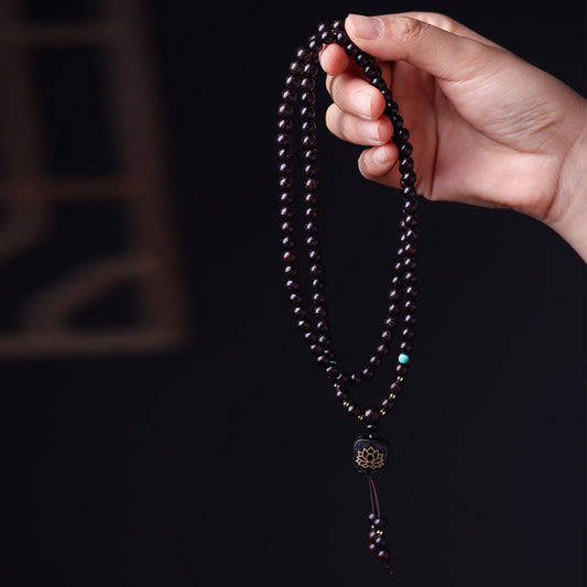 Hand holding 108 bead red sandalwood mala prayer beads with turquoise markers and gold lotus guru bead pendant against black background with blurred traditional lattice window
