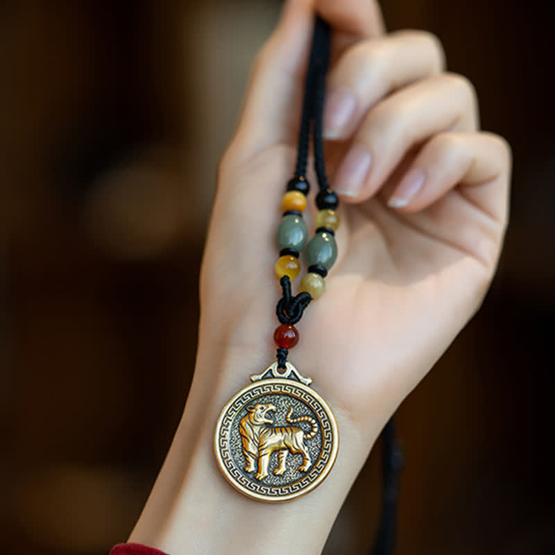 12 Chinese Zodiac Fortune Necklace Pendant – Tiger zodiac medallion pendant held in hand, antique gold brass with stippled background texture, roaring tiger relief, Greek key border, black cord with jade amber carnelian beads, traditional Chinese amulet necklace