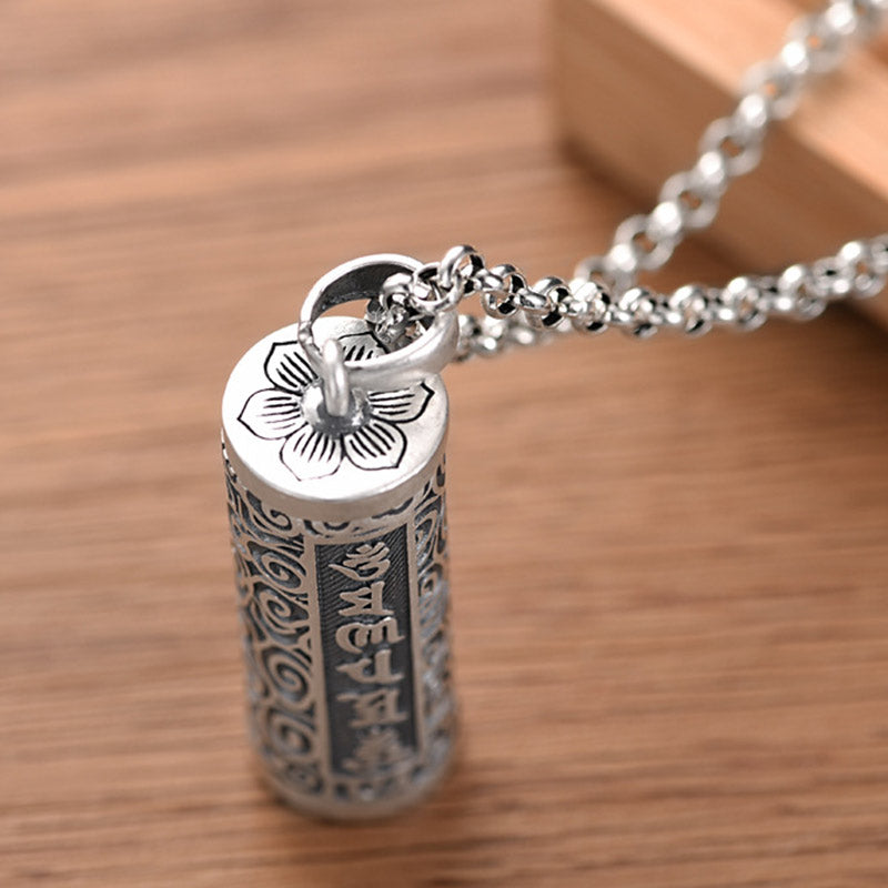 Om Mani Padme Hum Silver Protection Necklace – Close-up of silver cylindrical pendant with engraved lotus flower on top cap and vertical Tibetan script panels on cable chain against wood background